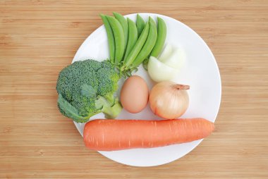 Ahşap tahta arka plan (brokoli, havuç, yumurta, soğan ve tatlı bezelye karşı beyaz tabakta taze sebze)