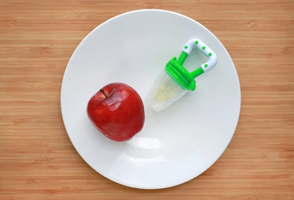 Baby\'s nibbler and red apple on white plate against wooden background. Organic baby food concept.