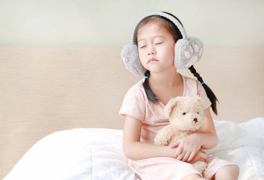Evde yatakta otururken kış earmuffs giyen sevimli küçük Asyalı çocuk kız ve oyuncak ayı sarılmak.