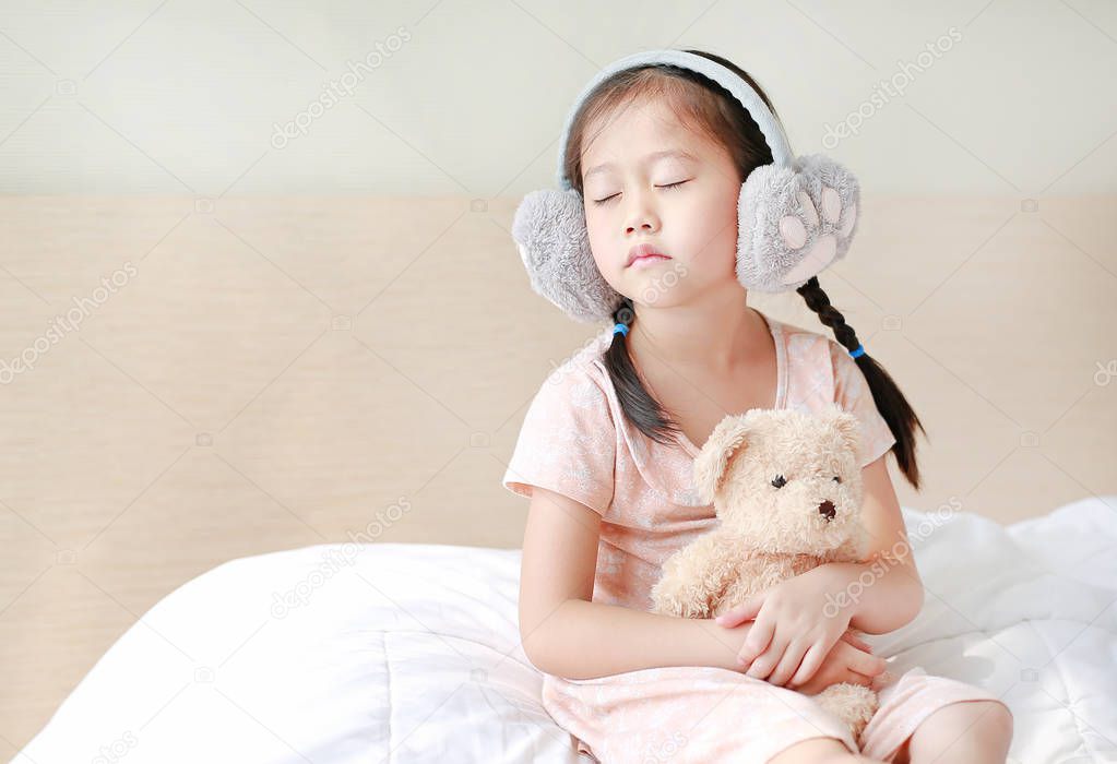 Adorable little Asian child girl wearing winter earmuffs and hugging teddy bear while sitting on the bed at home.