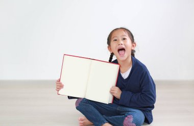 Asyalı küçük kız açık bir kitap gibi boş sayfayla gülüyor. Çocuk odada oturuyor ve elinde kitap tutuyor..