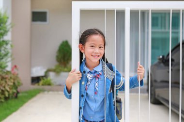Anaokulu üniformalı mutlu Asyalı kız sabah okula gitmeden önce evin çitlerinde..