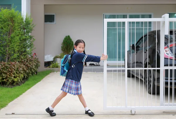 Cheerful asian little child girl in kindergarten uniform trying push and pull door fence of house to close or open before leave home to go to school at morning