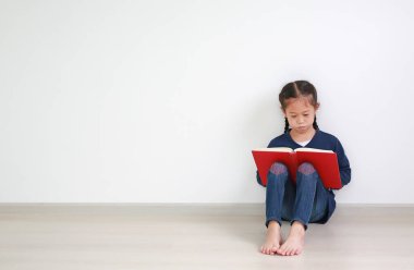 Portrait asian little kid girl in school uniform reading a book and sitting against white wall in the room with copy space. clipart