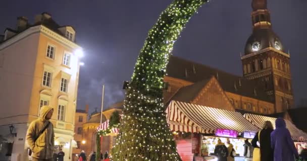 Riga, Letônia - 18 de dezembro de 2017: Pessoas caminhando perto do tradicional mercado de Natal na Praça Dome com a Catedral de Riga Dome. Marco famoso na noite de inverno na iluminação de Iluminações festivas — Vídeo de Stock