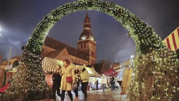 Riga, Letonia - 18 de diciembre de 2017: Gente caminando cerca del tradicional mercado de Navidad en la plaza de la cúpula con la catedral de la cúpula de Riga. Monumento famoso en la noche de invierno en iluminaciones festivas Iluminación — Vídeos de Stock
