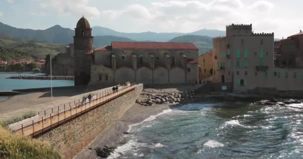 Collioure, França. Pessoas Turistas que caminham na costa perto da igreja de Nossa Senhora dos Anjos através da baía na ensolarada Primavera da Europa — Vídeo de Stock