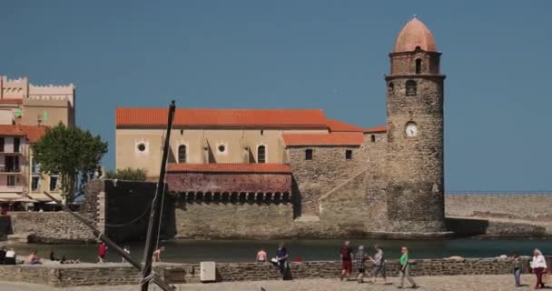 Collioure, França - 16 de maio de 2018: Turistas descansam e caminham na costa perto da igreja de Nossa Senhora dos Anjos do outro lado da baía na Primavera da Europa — Vídeo de Stock