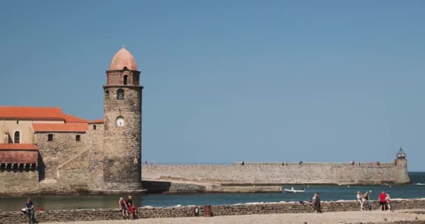 Collioure, Frankrijk - 16 mei 2018: Mensen Toeristen rusten en wandelen in de kust in de buurt van de kerk van Onze Lieve Vrouw van de Engelen aan de overkant van de baai in het voorjaar — Stockvideo