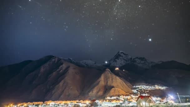 Stepantsminda, Georgia. Céu estrelado da noite com estrelas brilhantes sobre o pico do monte Kazbek coberto com neve. Famosa Igreja Gergeti In Night Lightning. Paisagem georgiana bonita — Vídeo de Stock