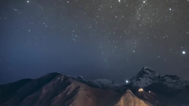 Stepantsminda, Georgien. Nächtlicher Sternenhimmel mit glühenden Sternen über dem Gipfel des Kazbek, der mit Schnee bedeckt ist. Berühmte Gergeti-Kirche in der Nacht Blitz. Schöne georgische Landschaft — Stockvideo