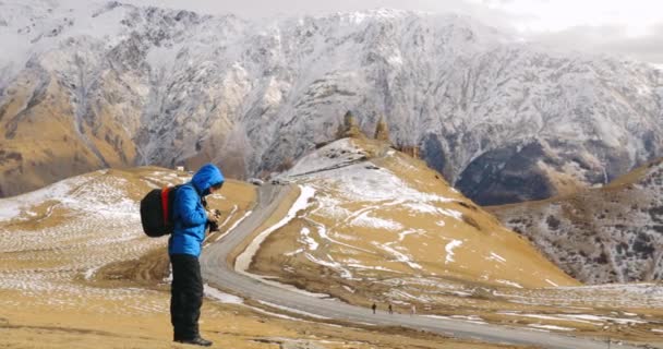 Stepantsminda, Gergeti, Georgia. Hombre Turista Mochilero Viajero Fotógrafo Tomando Fotos De Iglesia Santísima Trinidad - Tsminda Sameba. Hermoso paisaje georgiano a principios de invierno — Vídeos de Stock