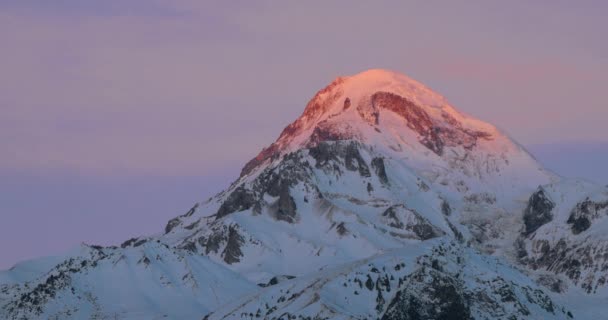 Stepantsminda, Gergeti, Geórgia. Monte Kazbek coberto de neve no nascer do sol de inverno. Morning Dawn Colored Top Of Mountain In Pink-Orange Colors (em inglês). Impressionante inverno paisagem da natureza georgiana — Vídeo de Stock