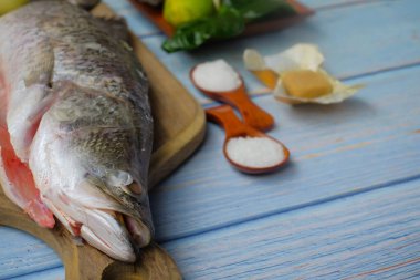 Bıçaklı, biberli, soğanlı, limonlu, kaffir limon yapraklı, zencefilli, sarımsaklı ve ağaç kesme tahtasının üzerinde yeşil limonlu Barramundi veya Asya deniz kabuğu balığı..