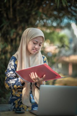 Başörtüsü takmış, not okuyan ve gümüş dizüstü bilgisayar kullanan İslami kız öğrencinin fotoğrafı.