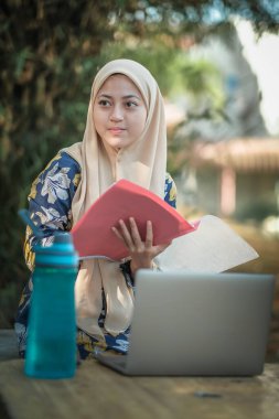 Başörtüsü takmış, not okuyan ve gümüş dizüstü bilgisayar kullanan İslami kız öğrencinin fotoğrafı.