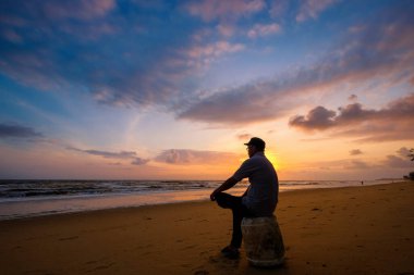 Tanımlanamayan adam güneş doğarken kumlu sahilde yumuşak mavi okyanus dalgalarıyla oturuyor.