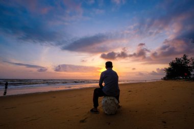 Tanımlanamayan adam güneş doğarken kumlu sahilde yumuşak mavi okyanus dalgalarıyla oturuyor.