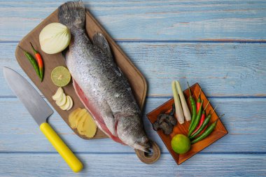 Bıçaklı, biberli, soğanlı, limonlu, zencefilli, sarımsaklı ve ağaç kesme tahtasının üzerindeki limonlu Barramundi veya Asya deniz kabuğu balığı..