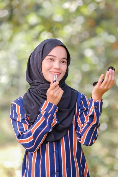 スマイリー美しいです若いですイスラム教徒女性身に着けていますHihab適用口紅保持手ミラー上のぼやけた背景 — ストック写真