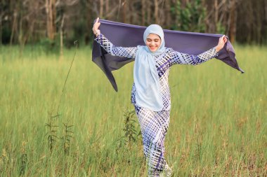 Elinde kumaş tutarken yeşil alanda poz veren şık tesettürlü genç ve mutlu bir mankenin portresi. Tarz sahibi Müslüman kadın tesettür moda tasviri.