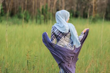 Elinde kumaş tutarken yeşil alanda poz veren şık tesettürlü genç ve mutlu bir mankenin portresi. Tarz sahibi Müslüman kadın tesettür moda tasviri.