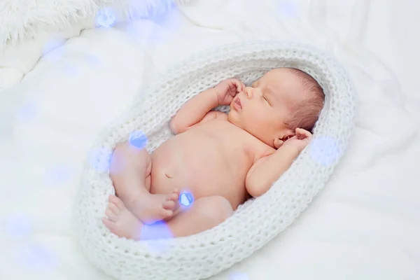 A cute adorable newborn baby wrapped in a white knitted cocoon sleeps with its legs bent in the Lotus position.  Blurred blue garlands ahead.