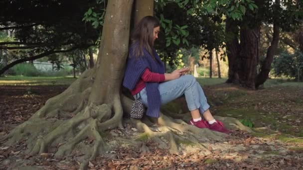 Dulce chica con un teléfono inteligente está sentado debajo de un árbol con poderosas raíces sinuosas — Vídeo de stock