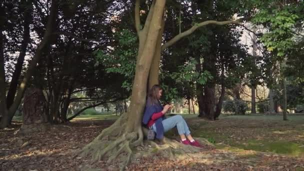 Süßes Mädchen mit Handy in der Hand sitzt unter einem großen Baum mit verdrehten Wurzeln, die draußen wachsen — Stockvideo