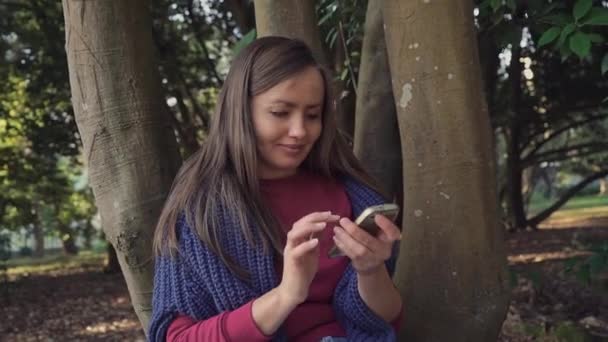 Tatlı kız o akıllı telefon kullanıyor ve gülümseyerek parkta ağacın yanında oturuyor — Stok video