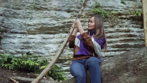 Улыбающаяся девушка в спортивной куртке сидит на дереве в горах и фотографирует по телефону — стоковое видео