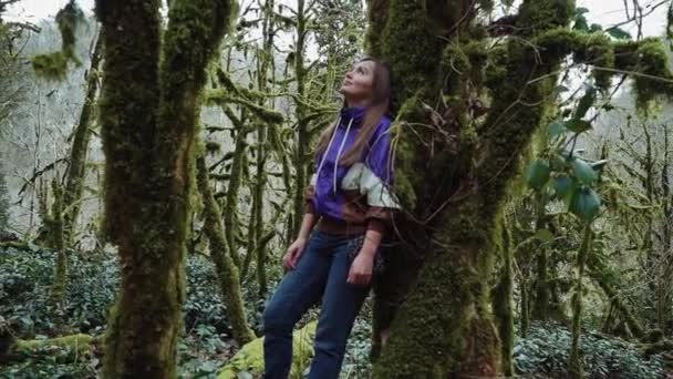 신비한 숲에서 이끼로 덮인 나무에 기대어 서있는 소녀 — 비디오