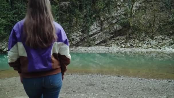 Menina com cabelos longos loiros em uma jaqueta e jeans chegando a um lago de montanha azul claro, vista das costas — Vídeo de Stock