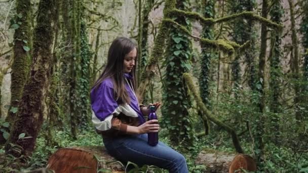 Девушка в спортивной куртке сидит на пне в лесу и откручивает крышку тепловой бутылки, чтобы выпить из нее . — стоковое видео