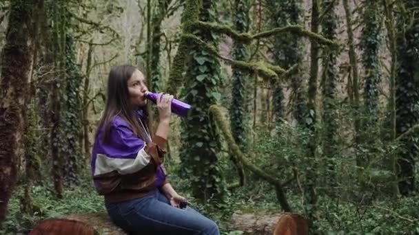 穿着运动夹克的女孩坐在森林的树桩上，她正在喝着一个彩色的热水瓶. — 图库视频影像