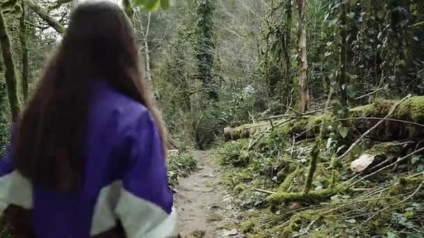 Menina de cabelos compridos em uma jaqueta de esportes vai em uma trilha florestal passado as árvores misteriosas cobertas com musgo verde . — Vídeo de Stock