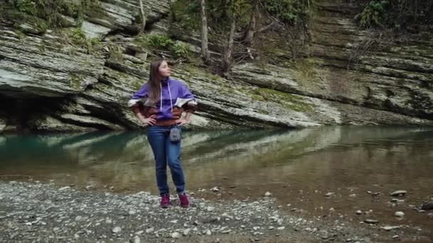 Красивая девушка стоит, положив руки на бедра на берегу чистой реки — стоковое видео