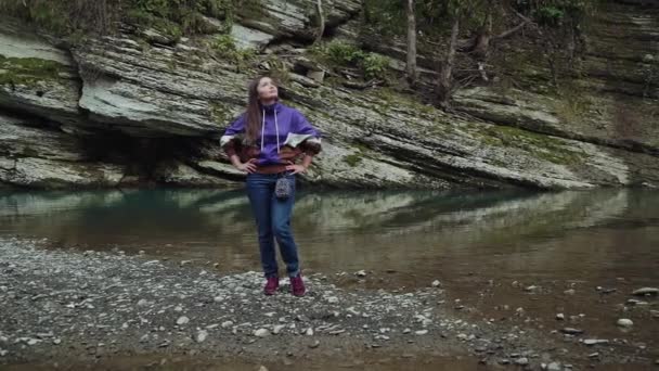 Chica feliz está de pie poniendo sus manos en las caderas junto al lago de la montaña y ella mira hacia arriba — Vídeo de stock