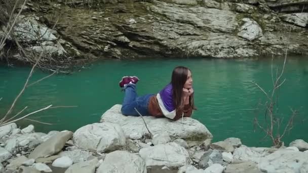 Uzun saçlı kız turkuaz bir dağ gölü yakınında büyük bir taş üzerinde yatıyor ve bacaklarını sallar. — Stok video