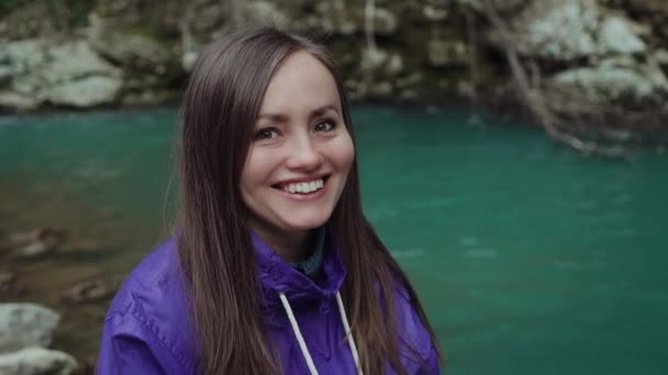 Édes boldog lány néz a kamerába, és mosolyog nagyjából a háttérben egy türkiz hegyi tó. — Stock videók