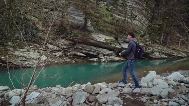 Высокий красивый парень с рюкзаком стоит на берегу чистой горной реки и бросает камни в воду . — стоковое видео