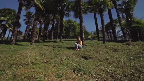 Mooi meisje in jeans en jasje zit op een groen gazon onder palmbomen — Stockvideo