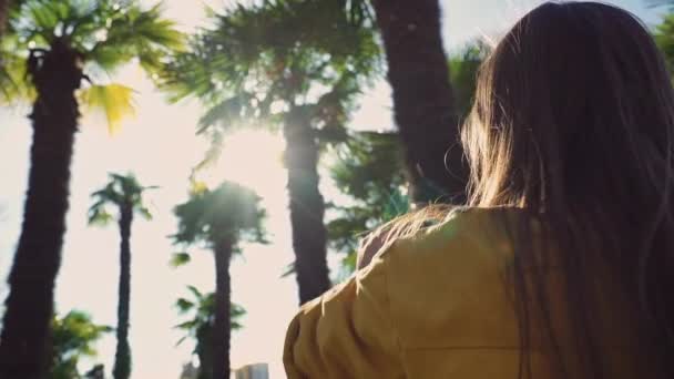 Hosszú hajú lány egy sárga kabátot lát gyönyörű kilátás pálmafák és elkezdi, hogy a képeket, hogy a smartphone — Stock videók