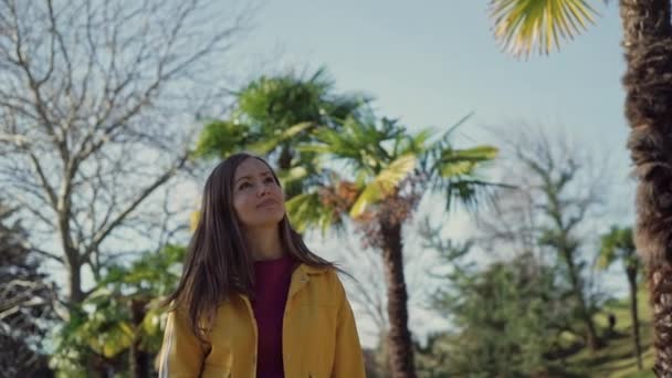 Hermosa chica feliz en una chaqueta amarilla camina por la avenida con palmeras en un día soleado — Vídeos de Stock