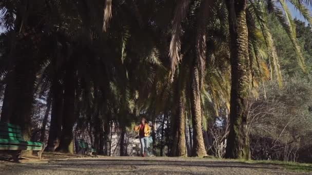 穿着牛仔裤和黄色夹克的快乐女孩带着棕榈树沿着小巷跑，有时跳起来，抓住树叶。慢动作 — 图库视频影像