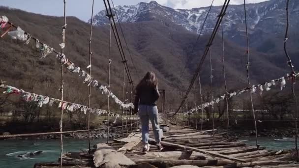 Chica se cuela a lo largo de un puente colgante en ruinas sobre un río limpio de montaña. Vista trasera — Vídeos de Stock