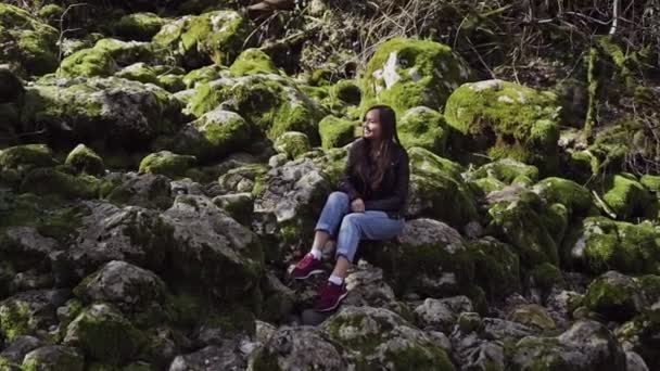 Dulce chica sonriente está sentado entre las piedras grandes dispersas que están cubiertas de musgo verde — Vídeos de Stock