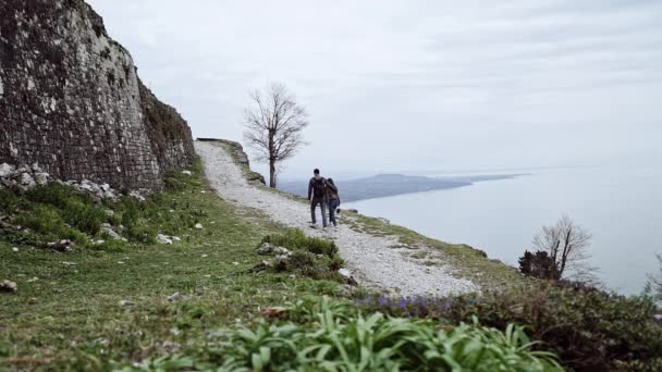 Пара влюбленных весело прогуляться по тропинке на краю скалы на фоне моря и гор, вид сзади — стоковое видео