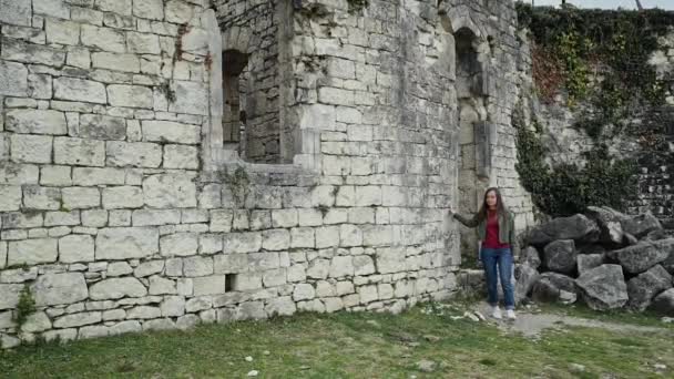 Дівчина в повсякденному одязі виходить зі старого замку, йде вздовж стіни і досліджує його — стокове відео