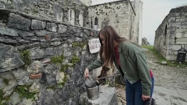 Mädchen trinkt aus einem Blechbecher an einem alten Steinbrunnen — Stockvideo